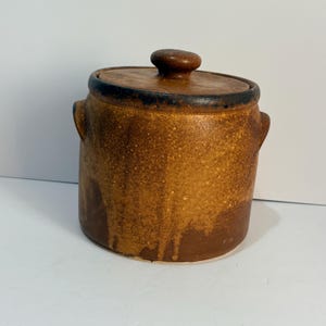 May include: A cylindrical brown ceramic jar with a matching lid and two small side handles. The exterior features a mottled, textured glaze with darker brown drip patterns running down the sides and a dark rim around the top edge.