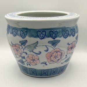 Puede incluir: Una maceta redonda de cerámica blanca con un diseño floral azul y rosa. La maceta tiene una abertura ancha y un borde azul decorativo en la parte superior e inferior. El patrón floral incluye flores rosas y hojas verdes.
