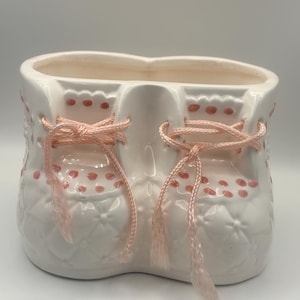 Könnte beinhalten: Ein weißer Keramik-Babyschuh-Pflanztopf mit rosa Akzenten. Der Pflanztopf hat rosa gepunktete Details und rosa Schnürsenkel. Der Schuh hat ein gestepptes Muster und ist für kleine Pflanzen oder Blumen gedacht. Ein charmantes Dekorationsstück.