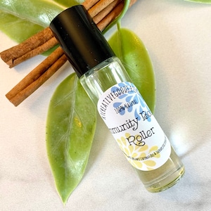 May include: A clear glass roller bottle with a black cap and a white label that reads "Creative Goods Est. 100% Natural Immunity Boost Roller" with a blue flower design. The bottle is sitting on a white surface with green leaves.