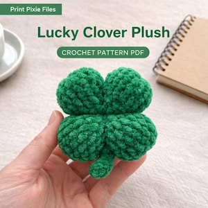 Puede incluir: Un trébol de cuatro hojas verde hecho a mano, de peluche, sostenido en una mano. El trébol está hecho de un hilo suave y texturizado. El texto "Lucky Clover Plush" y "CROCHET PATTERN PDF" se muestran encima del trébol.