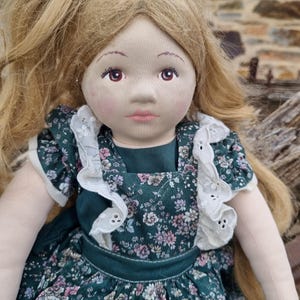 May include: A handcrafted cloth doll with long blonde hair and a pink bow. The doll features a painted face with rosy cheeks and a green floral dress with white lace trim. A charming example of handcrafted artistry.