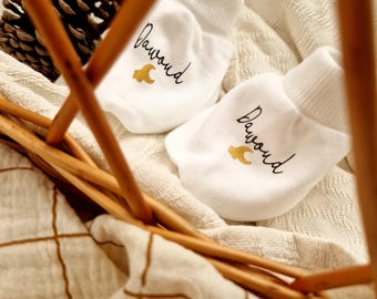 May include: Two white baby mittens with the name "Dawoud" in black script and a gold crescent moon design. The mittens are on a white blanket inside a wicker basket, with a pinecone visible.