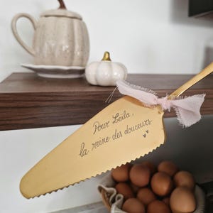 Puede incluir: Una paleta dorada para tartas con borde dentado y una cinta rosa atada cerca del mango. La paleta tiene el texto "Pour Leila, la reine des douceurs" escrito. Una cesta de huevos marrones está en primer plano.