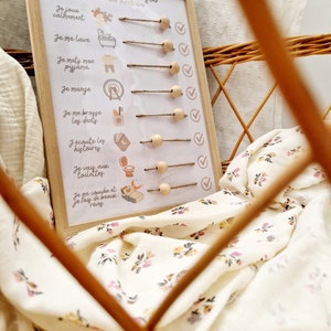 May include: A wooden-framed chart with the text "La routine du soir de Andr&eacute;a" and illustrated steps for a bedtime routine. The chart includes wooden pegs and check marks. The chart is in a wicker basket with a floral patterned fabric.