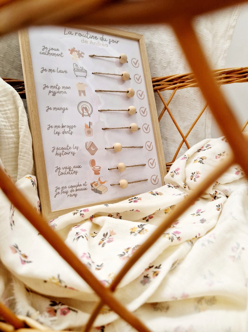 May include: A wooden-framed chart with the text "La routine du soir de Andr&eacute;a" and illustrated steps for a bedtime routine. The chart includes wooden pegs and check marks. The chart is in a wicker basket with a floral patterned fabric.