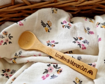 May include: A wooden spoon with a rounded bowl and a long handle. The handle is engraved with the text "La cuillère de Maman Ours" and a rainbow. The spoon rests on a white floral patterned cloth.
