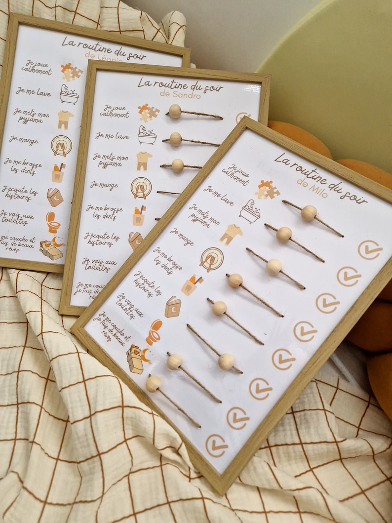May include: Three framed chore charts with the text "La routine du soir" and names. Each chart has illustrations and text in French, with wooden beads on strings to track tasks. The frames are light wood.