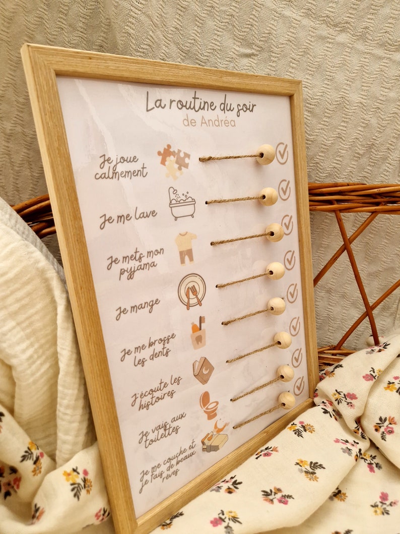 May include: A wooden framed chart with the text "La routine du soir de Andr&eacute;a" and illustrated steps for a bedtime routine. The chart includes text and icons for activities like playing, bathing, and brushing teeth. Wooden beads are attached to the chart.
