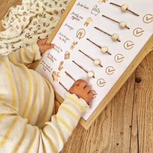 May include: A wooden-framed chart with the text "La routine du soir de Mia" and illustrated steps for a bedtime routine. The chart includes wooden beads on twine and checkmark icons. A child's arm in a yellow and white striped shirt is visible.