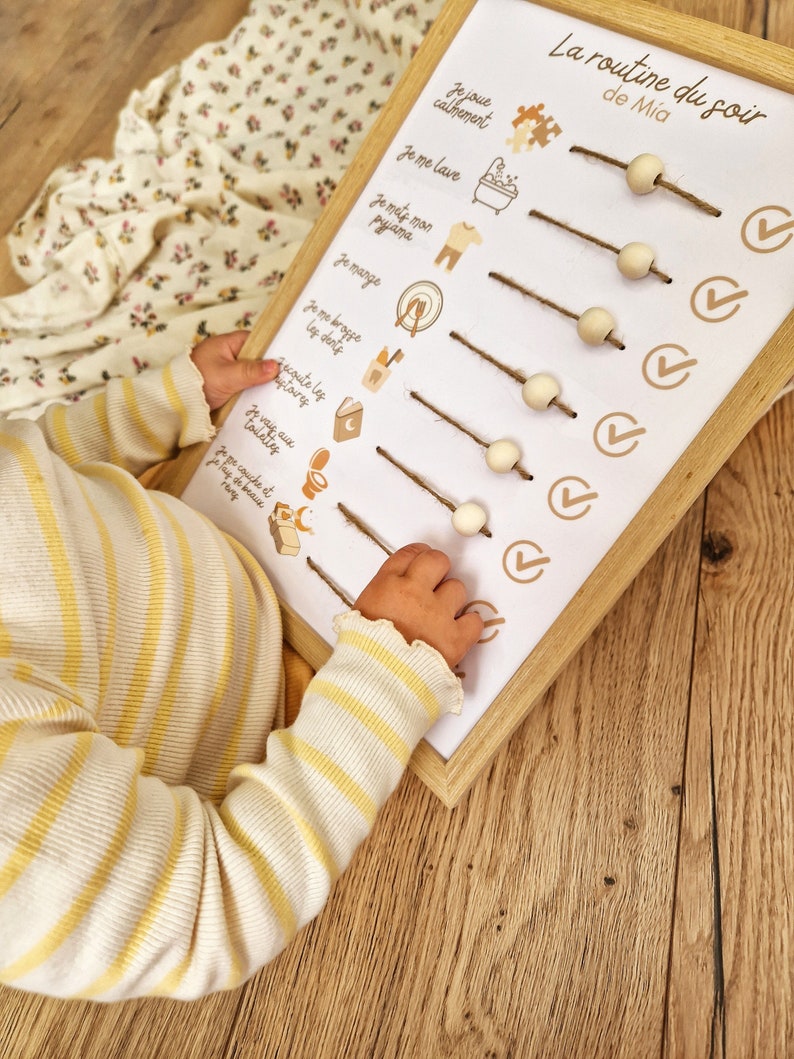May include: A wooden-framed chart with the text "La routine du soir de Mia" and illustrated steps for a bedtime routine. The chart includes wooden beads on twine and checkmark icons. A child's arm in a yellow and white striped shirt is visible.