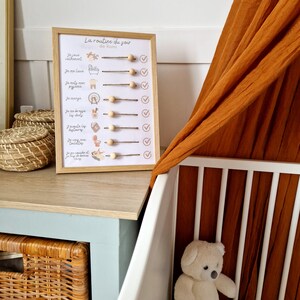 May include: A framed bedtime routine chart with the text "La routine du soir" and illustrations. The chart includes wooden beads and check marks. A white crib and a teddy bear are in the background.