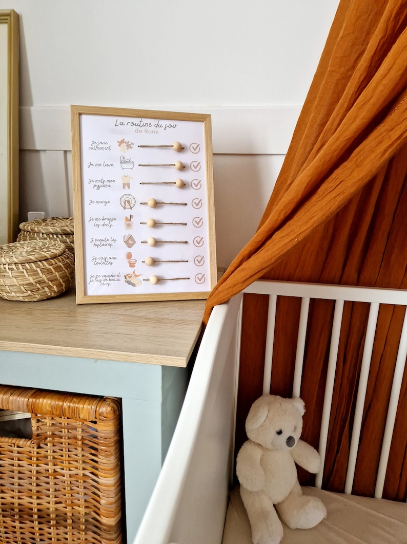 May include: A framed bedtime routine chart with the text "La routine du soir" and illustrations. The chart includes wooden beads and check marks. A white crib and a teddy bear are in the background.