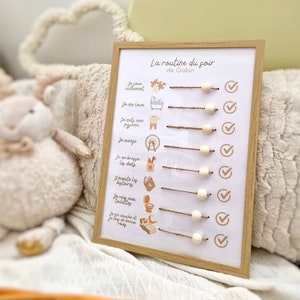 May include: A framed, white printed chart with the text "La routine du soir de Gabin" in French. The chart has illustrated icons and text describing a bedtime routine, with wooden beads and twine. The frame is light brown.
