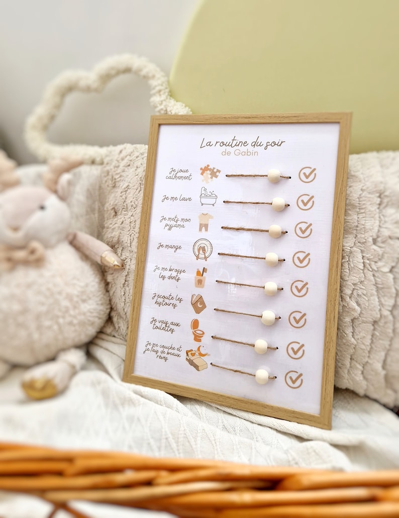 May include: A framed, white printed chart with the text "La routine du soir de Gabin" in French. The chart has illustrated icons and text describing a bedtime routine, with wooden beads and twine. The frame is light brown.