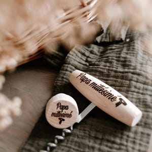 Könnte beinhalten: Ein hölzernes Korkenzieher- und Flaschenverschluss-Set. Der Korkenziehergriff und der Verschluss sind aus hellem Holz mit den Worten "Papa millésimé" graviert. Das Set liegt auf einem grauen Tuch.