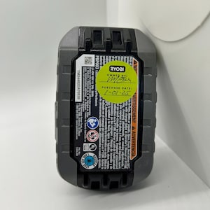 Pu&ograve; includere: Una batteria agli ioni di litio Ryobi PBP003 da 18 V grigia e nera. La batteria ha un adesivo con la scritta "OWNED BY" e "PURCHASE DATE: 1-01-25". La batteria ha anche etichette di avvertimento e informazioni di sicurezza stampate.
