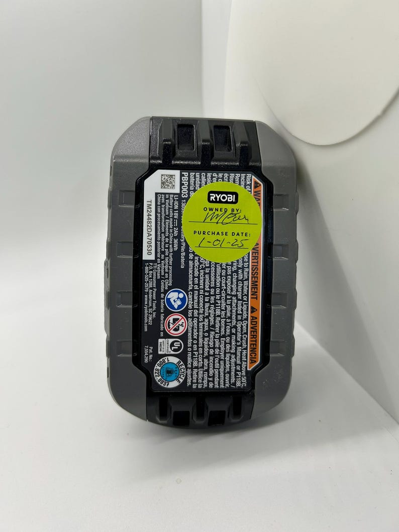 Pu&ograve; includere: Una batteria agli ioni di litio Ryobi PBP003 da 18 V grigia e nera. La batteria ha un adesivo con la scritta "OWNED BY" e "PURCHASE DATE: 1-01-25". La batteria ha anche etichette di avvertimento e informazioni di sicurezza stampate.