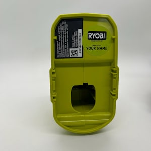 Pu&ograve; includere: Un pacco batteria Ryobi verde lime con forma rettangolare. Il pacco ha un adesivo con la scritta "PROPERTY OF YOUR NAME" e "RYOBI". Il pacco ha anche un adesivo con testo in inglese, francese e spagnolo.