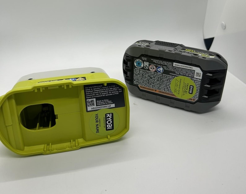 Pu&ograve; includere: Due componenti della batteria per utensili elettrici Ryobi da 18 V. Uno &egrave; una base caricatore verde lime e bianca con il testo "YOUR NAME" e "RYOBI". L'altro &egrave; un pacco batteria nero e grigio con un adesivo verde e varie etichette.