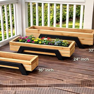 May include: A trio of wooden rectangular planters. The planters are constructed from light-toned wood and feature black metal supports. The largest planter is 81 cm long, the medium is 61 cm, and the smallest is 41 cm. The medium planter is filled with colorful flowers.