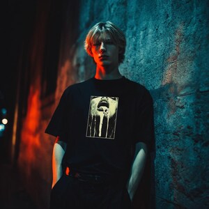 May include: Black t-shirt with a graphic print of a mouth with white drips. The person is standing in front of a textured blue wall, with red and orange lighting.