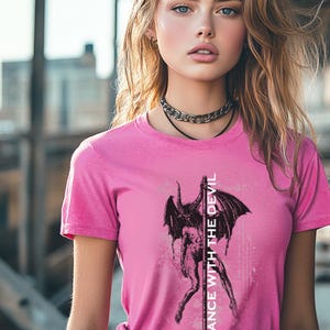 Könnte beinhalten: Pinkes T-Shirt mit einem schwarz-weißen Grafikdruck eines Dämons mit Flügeln und dem Text "Dance with the Devil", der vertikal auf der Vorderseite aufgedruckt ist.