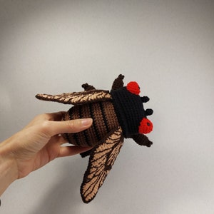 May include: A crocheted plush toy of a cicada with brown and black stripes, red eyes, and beige wings.