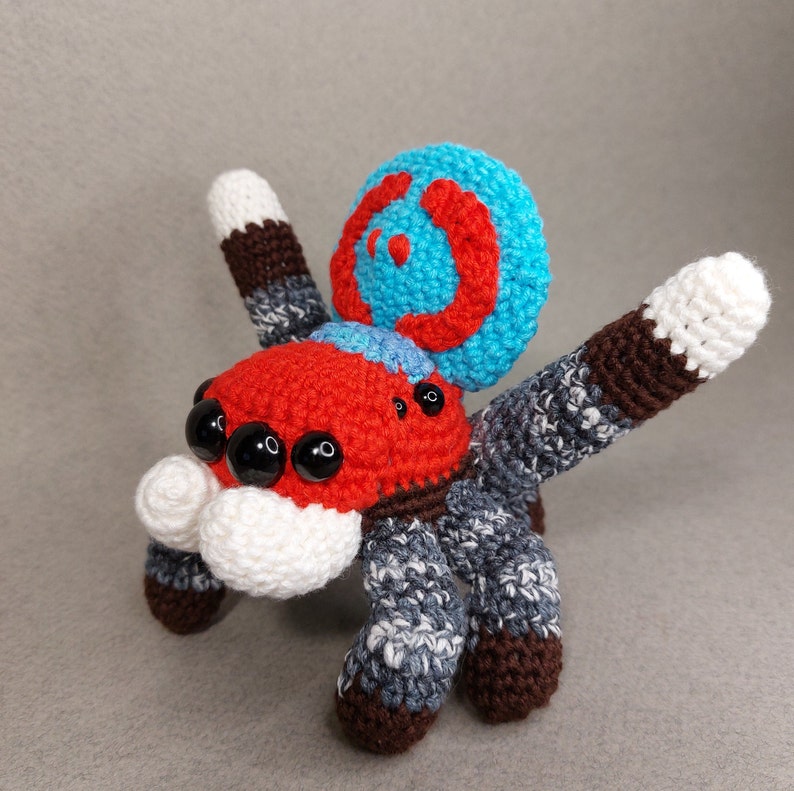Puede incluir: Un juguete de ara&ntilde;a de crochet con un cuerpo rojo, una cara blanca, ojos negros y una espalda con un patr&oacute;n azul y rojo.