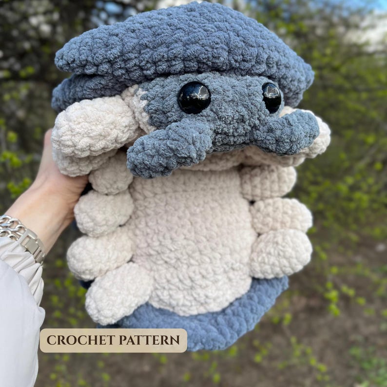 Crocheted plush roly poly pill bug in blue and cream blanket yarn, held outdoors in a hand, with large black eyes and layered body segments.