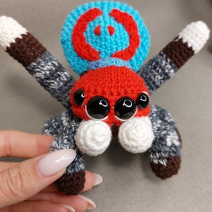 Puede incluir: Un juguete de ara&ntilde;a saltarina de crochet con un cuerpo rojo, una cabeza con estampado azul y rojo y ojos blancos. La ara&ntilde;a tiene ocho patas con rayas marrones y grises.