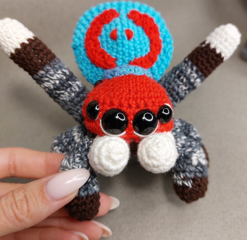 Puede incluir: Un juguete de ara&ntilde;a saltarina de crochet con un cuerpo rojo, una cabeza con estampado azul y rojo y ojos blancos. La ara&ntilde;a tiene ocho patas con rayas marrones y grises.