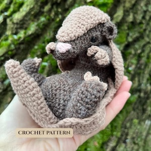 Puede incluir: Un pequeño juguete de armadillo tejido a ganchillo a mano con cuerpo marrón oscuro, caparazón color canela claro y una pequeña nariz rosa. El armadillo está en posición enroscada con puntadas texturizadas. Una etiqueta dice CROCHET PATTERN.