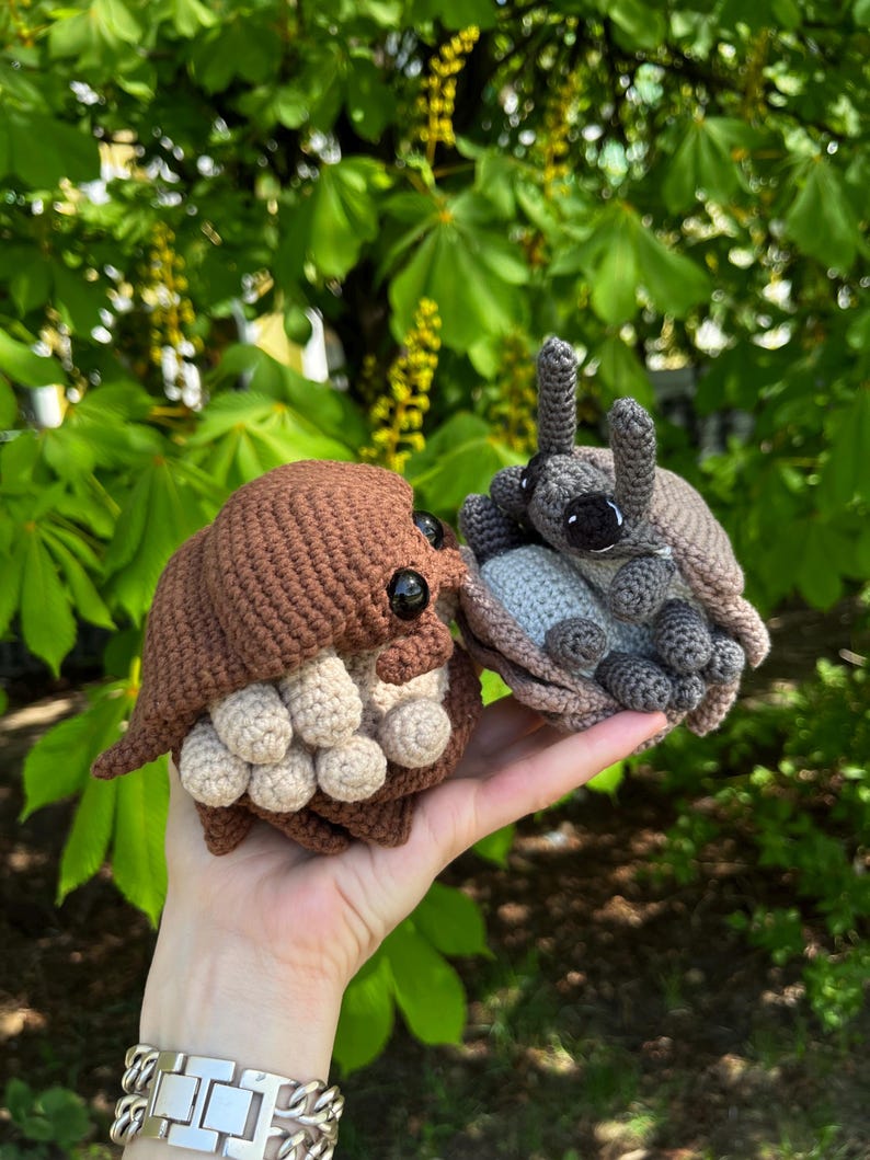 May include: Two small, crocheted woodlouse figures. One is brown with light beige underside segments and black bead eyes. The other is grey with a light grey underside, featuring two prominent antennae and black bead eyes. Both have rounded, segmented shells.