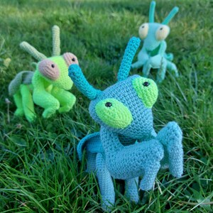 May include: Three crocheted green and blue mantis toys on a green grassy background. The mantises have large, round eyes and long, thin legs.