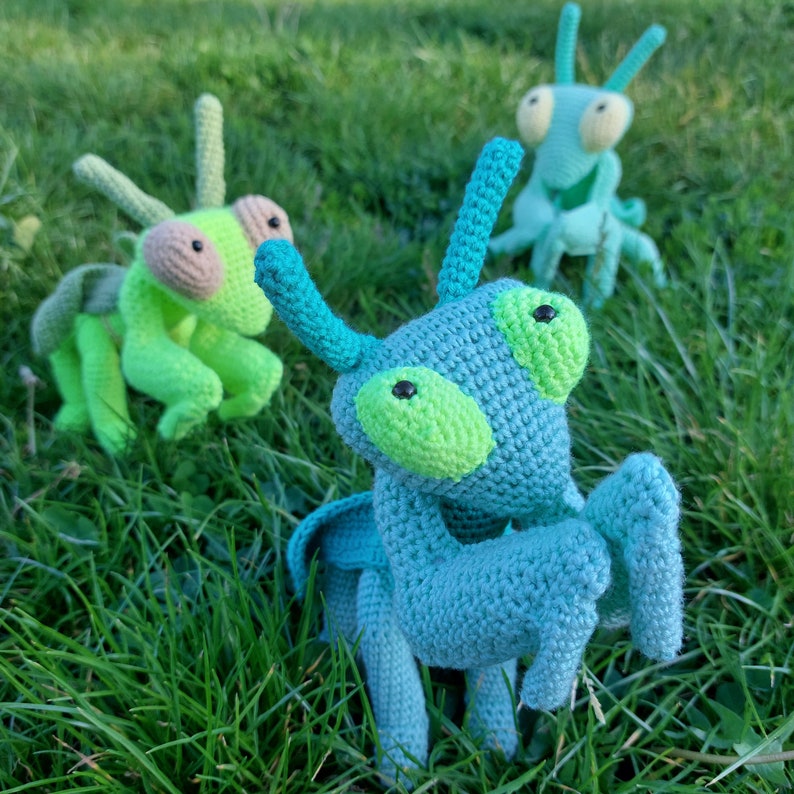 May include: Three crocheted green and blue mantis toys on a green grassy background. The mantises have large, round eyes and long, thin legs.