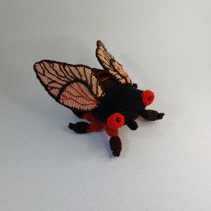 May include: A crocheted cicada toy with brown and orange wings, black body, and red eyes.
