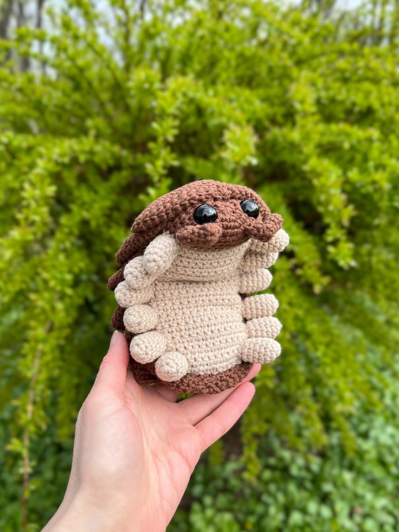 May include: A crocheted pill bug plush toy featuring a dark brown segmented shell and a light beige underside. The creature has two large, round, black plastic eyes positioned on the front of its head and multiple small, rounded legs along its sides.