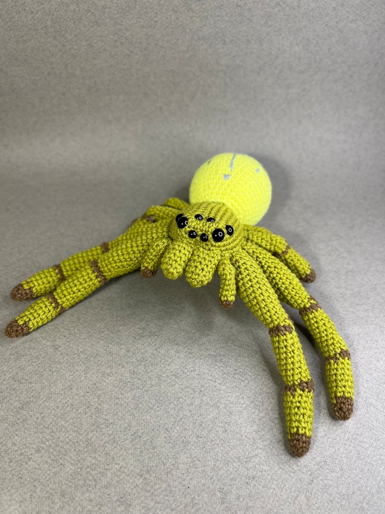May include: A crocheted green spider with brown accents and eight legs. The spider has a yellow head with black eyes.