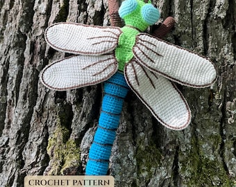 May include: A handcrafted crochet dragonfly toy featuring a bright green head with large light blue eyes, a long blue segmented body, and four expansive off-white wings with brown stitched details. The text CROCHET PATTERN is visible.