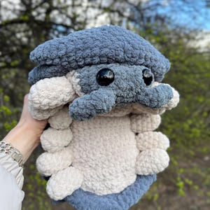 May include: A plush toy shaped like an isopod, crafted from soft, textured yarn. The creature features a dark blue segmented shell and head, with a light cream-coloured underside and multiple rounded legs. It has two large, round, black eyes.