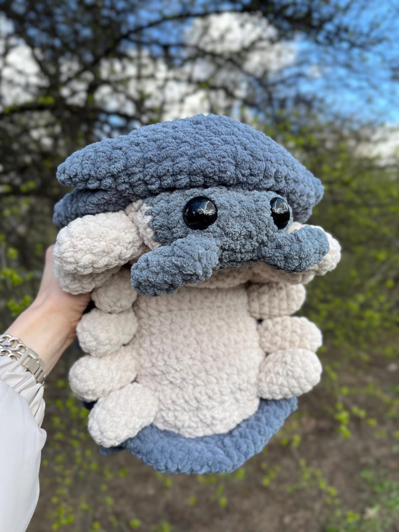 May include: A plush toy shaped like an isopod, crafted from soft, textured yarn. The creature features a dark blue segmented shell and head, with a light cream-coloured underside and multiple rounded legs. It has two large, round, black eyes.