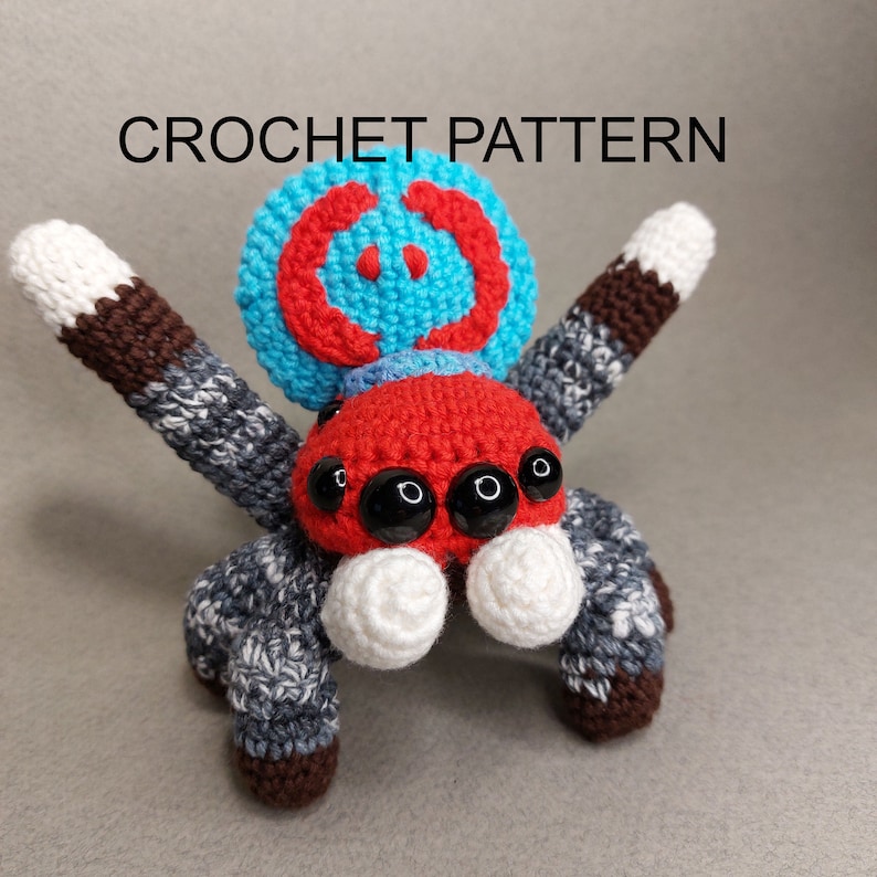 Puede incluir: Un juguete de ara&ntilde;a de crochet con un cuerpo rojo, una cabeza con estampado azul y rojo y ojos negros. La ara&ntilde;a tiene ocho patas con rayas marrones, grises y blancas.