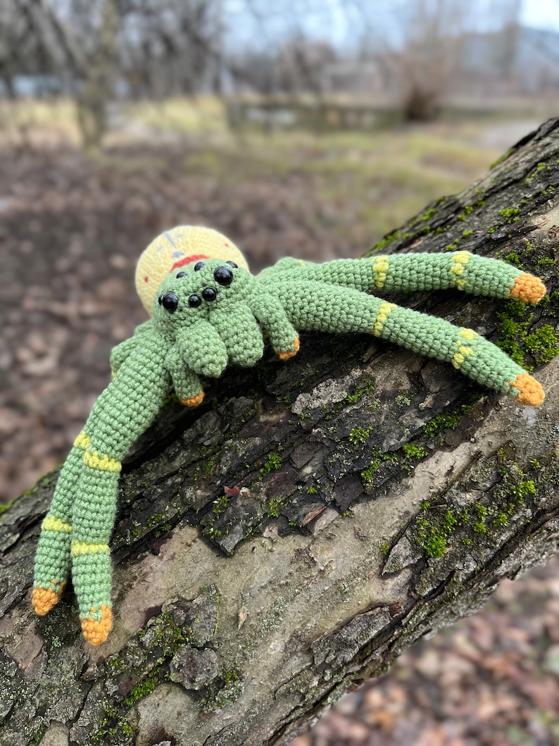 May include: A crocheted green spider with yellow accents and black eyes. The spider is sitting on a tree branch.