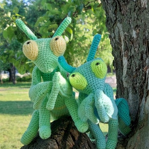 May include: Two crocheted green and blue mantis toys. The mantis on the left is green with yellow eyes and the mantis on the right is blue with green eyes.