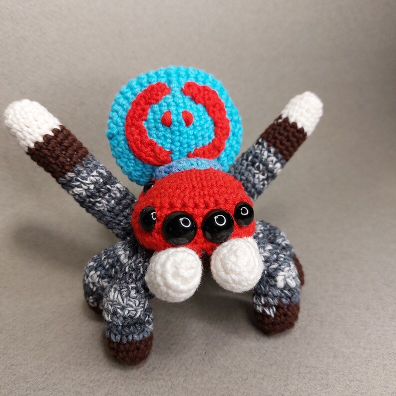 Puede incluir: Un juguete de ara&ntilde;a de crochet con un cuerpo rojo, ojos negros y una cabeza con un patr&oacute;n azul y rojo. La ara&ntilde;a tiene ocho patas, cada una con un patr&oacute;n de color diferente.