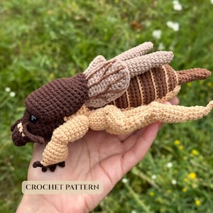 May include: A crocheted insect toy featuring a dark brown head, light beige legs, and a tan and brown striped body with textured wings. The toy has a small black eye and a label at the bottom that clearly displays the text CROCHET PATTERN.