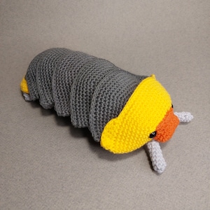 May include: A grey and yellow crocheted plush toy shaped like a pill bug. The toy has a yellow head with orange accents and black eyes.