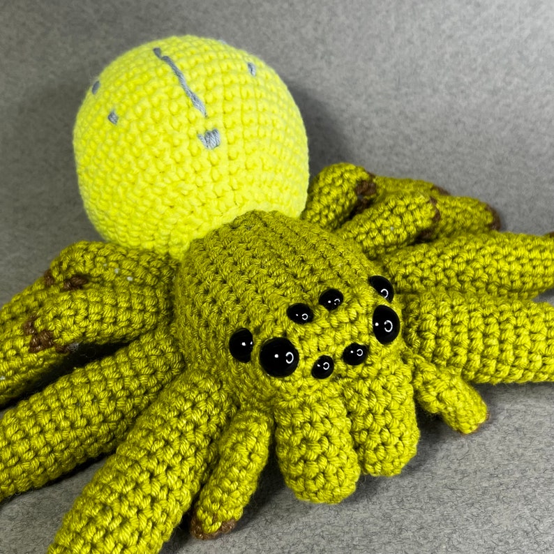 May include: A crocheted green spider with black eyes and a yellow head. The spider has eight legs and is made of yarn.