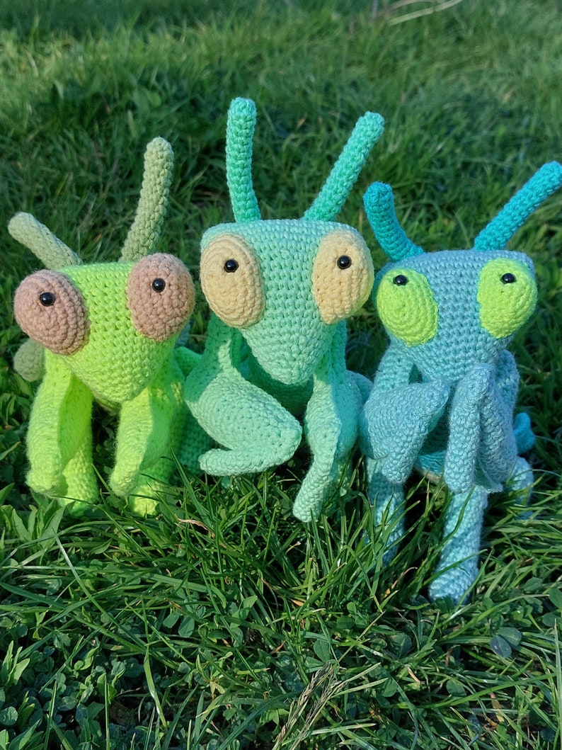 May include: Three crocheted mantis toys in different shades of green. The mantis on the left is a bright lime green, the middle mantis is a light green, and the mantis on the right is a teal blue.
